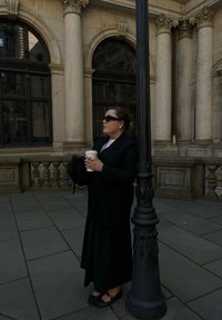 Juodas per didelis švarkas ant baltos palaidinės, ilga juoda sijonas, laikanti kavos puodelį, stovinti šalia lampo su klasikinių architektūrinių detalių.