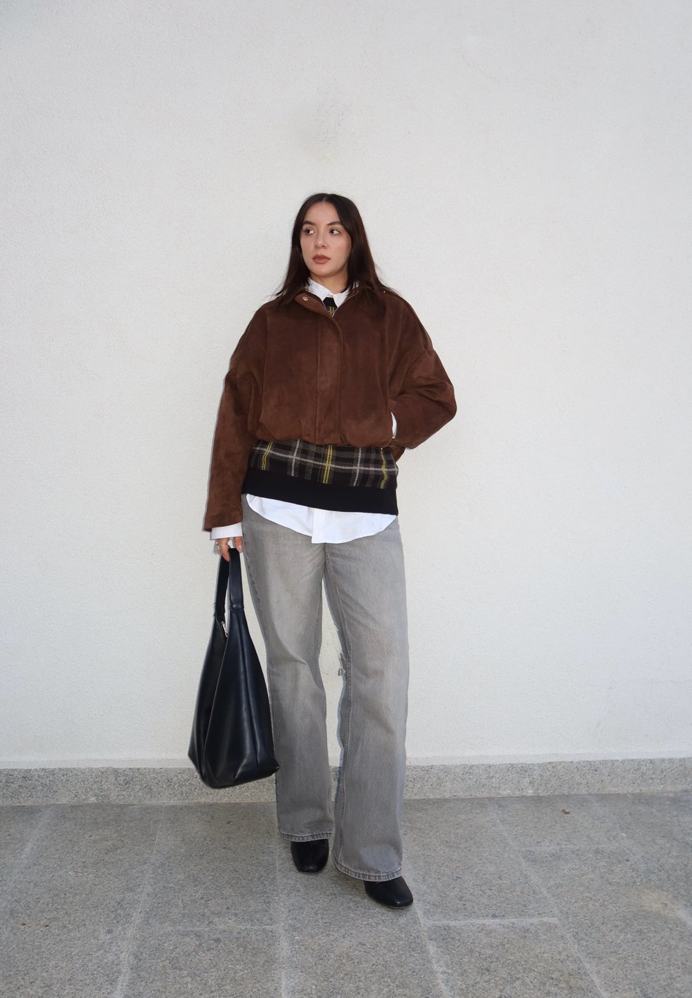 Veste bomber en daim marron portée sur une chemise blanche, pull à carreaux gris, pantalon gris à jambes larges, sac noir et bottines noires à talons.