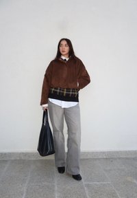 Veste bomber en daim marron portée sur une chemise blanche, pull à carreaux gris, pantalon gris à jambes larges, sac noir et bottines noires à talons.