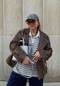 Καφέ δερμάτινο μπουφάν πάνω από ένα ριγέ πόλο μπλουζάκι, μπλε παντελόνι, κρατώντας μια μικρή ασημένια τσάντα. Τα αξεσουάρ περιλαμβάνουν γυαλιά ηλίου και ένα χοντρό κολιέ.