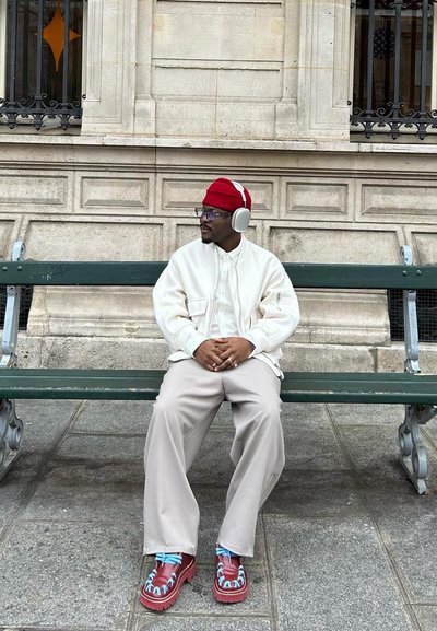 Chaqueta blanca con cremallera, camisa crema, pantalones amplios beige, gorro rojo, grandes auriculares grises y botas rojas con patrón y cordones azules.