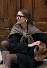 Μαύρο παλτό ριγμένο στους ώμους, καφέ πλεκτό κασκόλ δεμένο μπροστά, ασημένια σκουλαρίκια, μαύρα γυαλιά και κρατώντας μια καφέ δερμάτινη τσάντα.