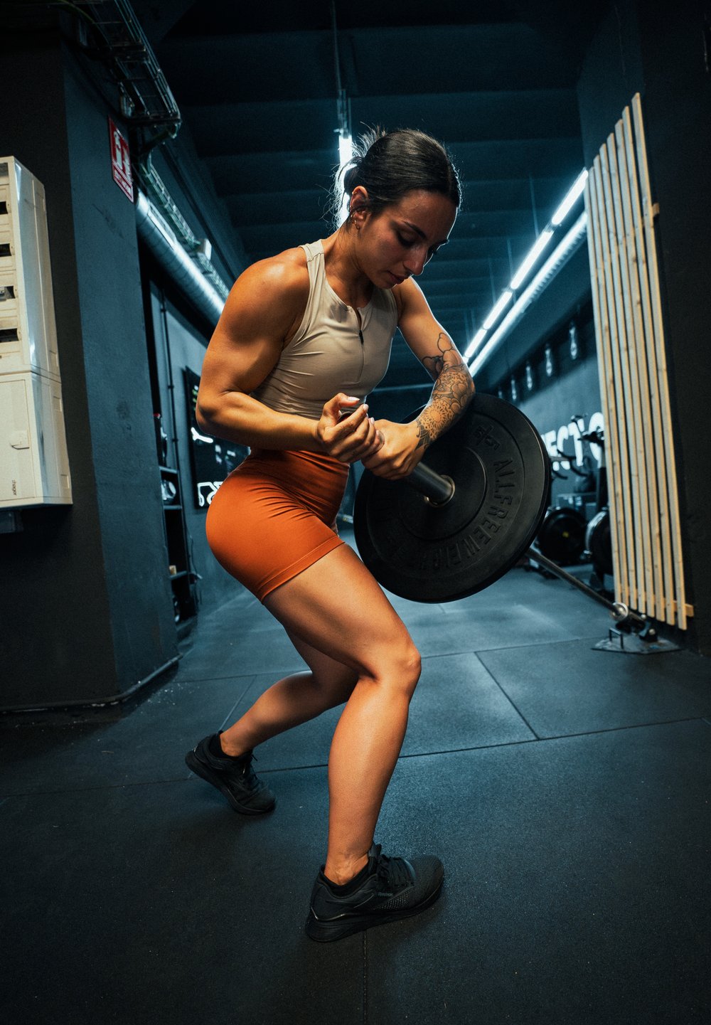 Athlète soulevant une barre noire sur le sol de la salle de sport ; portant un haut beige et un short orange. Jambes musclées et chaussures de sport noires visibles.