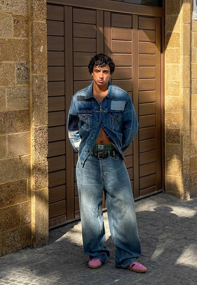 Chaqueta de mezclilla con tonos azules variados, vaqueros azules sueltos, cinturón verde y zapatos de lona rosas; de pie contra una pared de piedra texturizada.