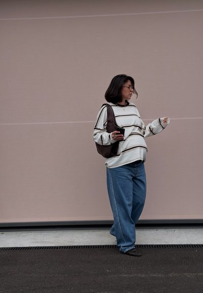 Mujer con cabello oscuro hasta los hombros, con gafas, suéter a rayas y vaqueros holgados, sosteniendo una taza y apoyada contra una pared beige.
