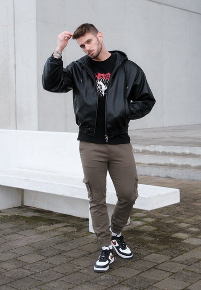 Chaqueta bomber de cuero negra, camiseta gráfica negra, pantalones cargo verde oliva, zapatillas negras y blancas, de pie contra un fondo gris claro.