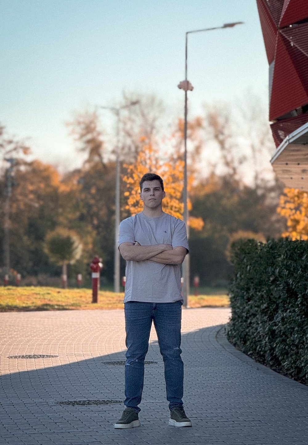 A person wearing a light grey t-shirt and blue jeans stands on a paved path with green foliage and autumn trees in the background.