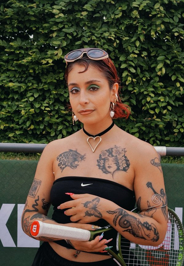 Ung kvinde med farverige tatoveringer og hjertehalskæde holder en tennisketcher og står foran en grøn, bladet baggrund.