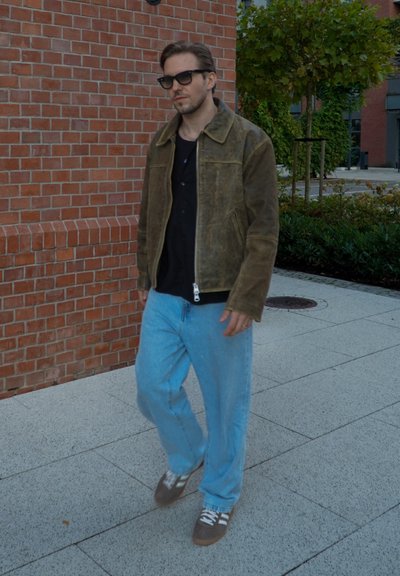 Hombre con gafas de sol caminando por la acera junto a una pared de ladrillo, vistiendo una chaqueta marrón, camiseta negra, jeans de color azul claro y zapatillas grises.