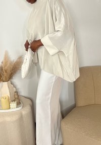 Top din satin bej cu mâneci largi, asortat cu pantaloni albi. Prezintă o poșetă texturată albă și vase neutre decorative pe o masă.