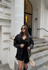 Giacca oversize in pelle nera su un vestito camicia di pizzo nero, abbinata a una borsa beige in tessuto, su scalini di pietra davanti a un edificio bianco.