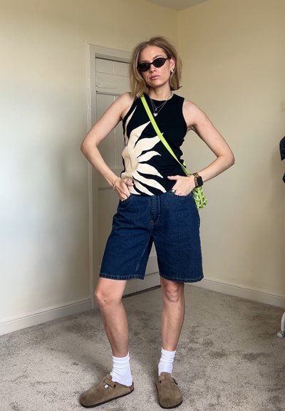 Top sin mangas negro con un diseño de sol crema, pantalones cortos de mezclilla, calcetas blancas y zuecos marrones. Un bolso cruzado verde añade contraste.
