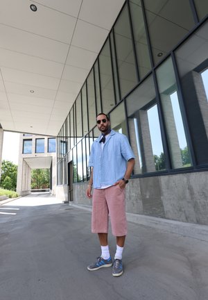 Camisa de manga corta de textura azul claro, pantalones cortos de pana rosa, zapatillas grises con acentos azules, de pie al aire libre cerca de grandes ventanales de cristal.
