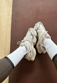 Pieds portant des baskets blanches épaisses et des chaussettes blanches côtelées reposant sur un tapis de gym marron avec un sol en bois clair à proximité.