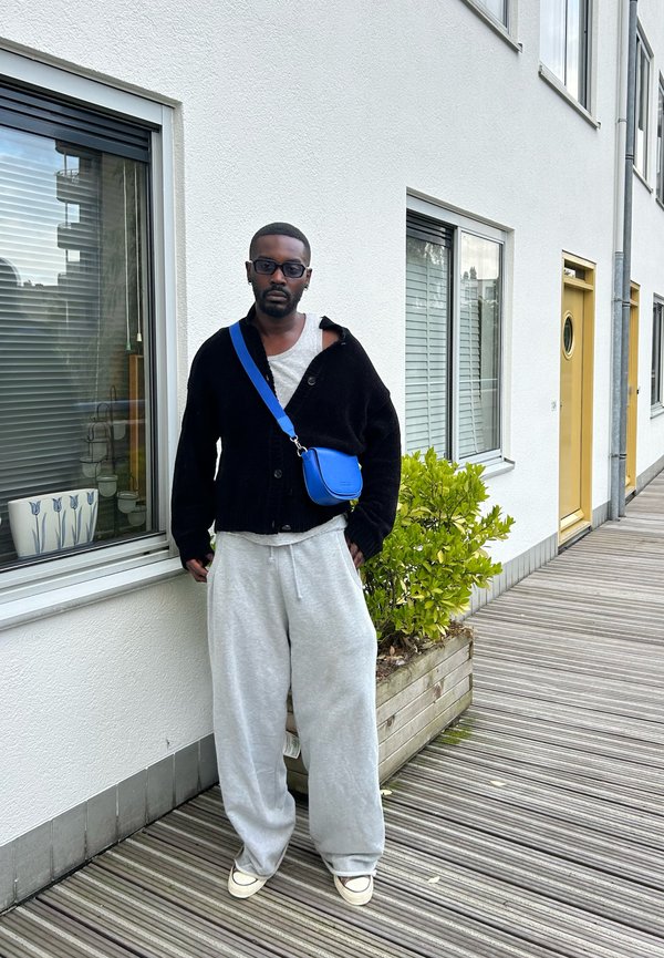 Cardigan nero su una maglietta grigia, pantaloni felpa grigi e scarpe nere. Borsa a tracolla blu. In piedi accanto a un edificio con finestre e una porta gialla.