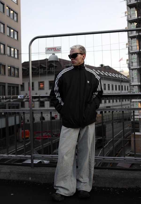 Negro chaqueta deportiva con tres rayas blancas y logo; vaqueros de pierna ancha azul claro; gafas de sol oscuras. El fondo incluye una valla de construcción y edificios.
