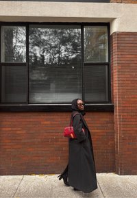 Cappotto lungo nero sopra un abito scuro, abbinato a una borsa rossa. Sullo sfondo, un muro di mattoni e grandi finestre con persiane.