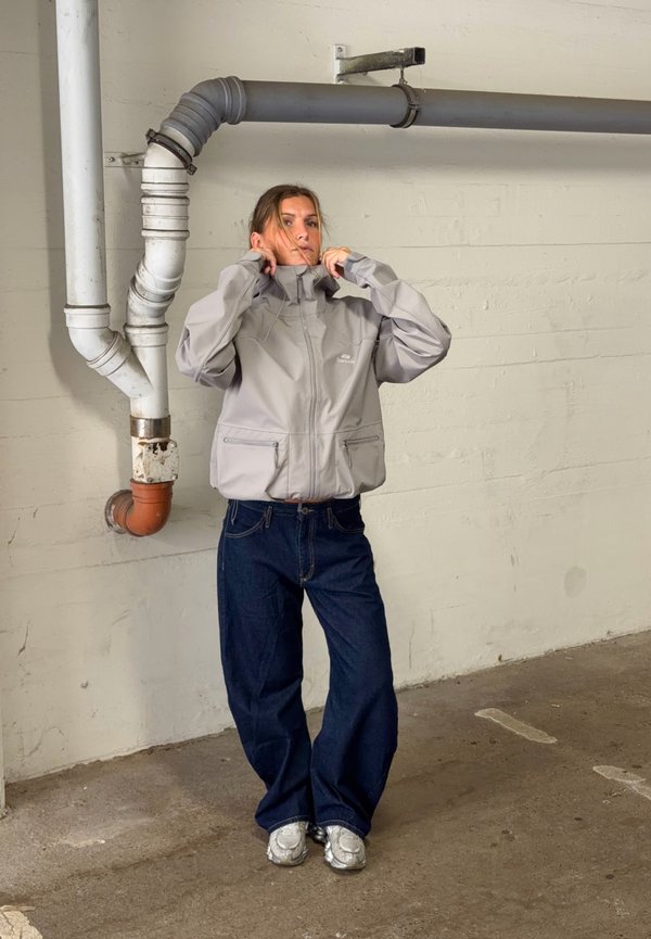 Femme ajuste le col d'une veste gris clair, portant un jean large bleu foncé et des baskets argentées, debout près des tuyaux muraux à l'intérieur.