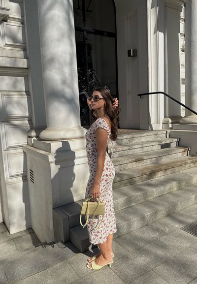 Vestido blanco con patrón floral rojo, mangas cortas y detalles fruncidos. Sandalias de tacón cuadrado amarillas y un pequeño bolso amarillo. Escaleras de piedra y columnas.