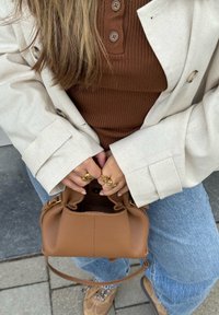 Brun læderhåndtaske med en snøret åbning, parret med en beige blazer over en brun ribbet top. Guld smykker på hænderne; denim jeans synlige.