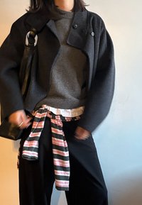 Personne portant un manteau gris foncé, un pull gris, un pantalon noir et portant un sac à bandoulière noir, avec une chemise rayée nouée autour de la taille.