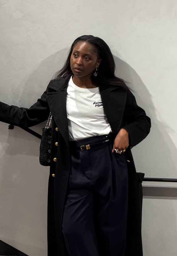 Mujer con cabello largo y oscuro, con un abrigo negro, camiseta blanca y pantalones oscuros, apoyada contra una pared gris claro y una barandilla.