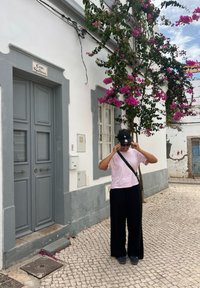 Siv door s dvoma panelmi a okrúhlou kľučkou, biela stena s ružovou bougainvilleou, kamenná cesta, osoba v ružovej košeli a čiernych nohaviciach.
