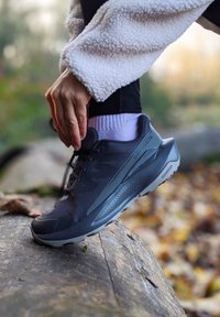 Shoe desportivo preto com parte superior em malha texturada, sola em borracha e biqueira reforçada. Acompanhado de uma meia rosa e leggings pretas sobre uma superfície roca.