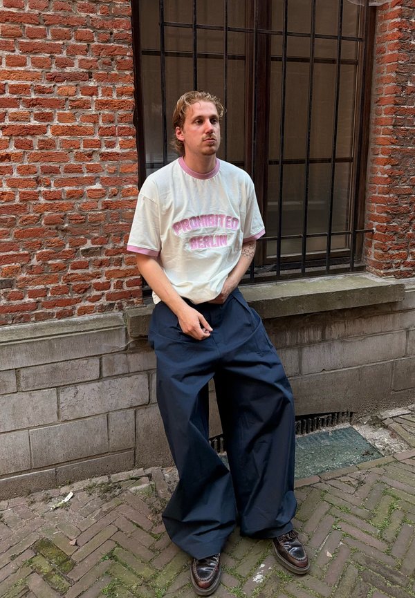 Homme aux cheveux châtain clair portant un t-shirt blanc avec un texte rose et un pantalon bleu foncé trop grand, appuyé contre un mur en briques sous une fenêtre grillagée.
