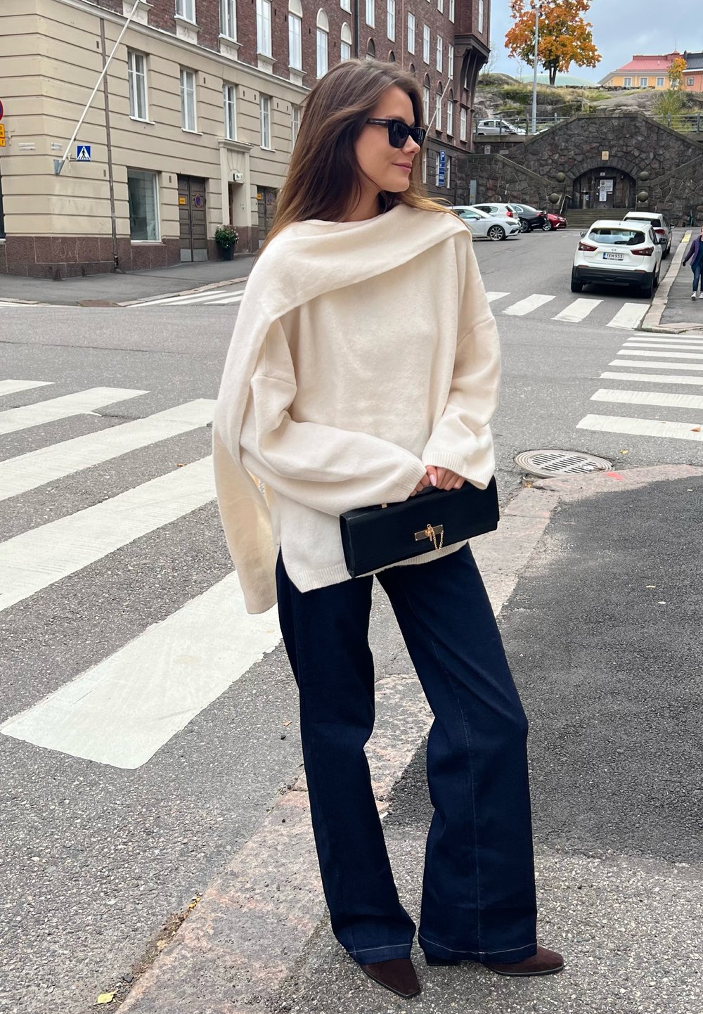 Mulher com um suéter oversized creme e calças largas escuras, segurando uma carteira preta, parada perto de uma faixa de pedestres numa rua da cidade.