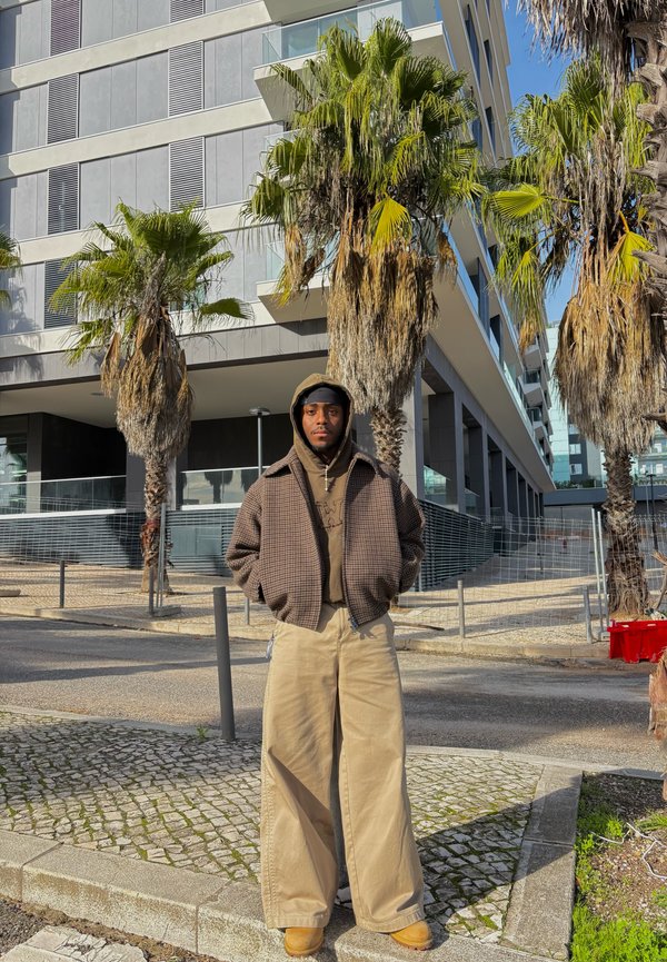 Getextureerd, bruin geruit jack over een beige hoodie, gecombineerd met wijde beige broek en tan laarzen. Op de achtergrond palmbomen en moderne gebouwen.