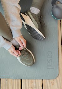 Chaussures de sport vertes avec des accents gris et des semelles texturées, attachées sur un tapis de yoga, avec une kettlebell à proximité. Sweat-shirt gris décontracté et leggings.
