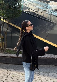 Crna prevelika jakna s ovratnikom u stilu šala, traperice i crna torbica preko tijela. Duga pletenica, nosi tamne sunčane naočale.