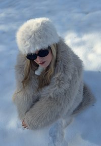 Vale karusnahast mantel ja müts heledas beežis; paks, pehme tekstuur. Mustad päikeseprillid. Taust: valge lumega kaetud pind, muid elemente ei ole.