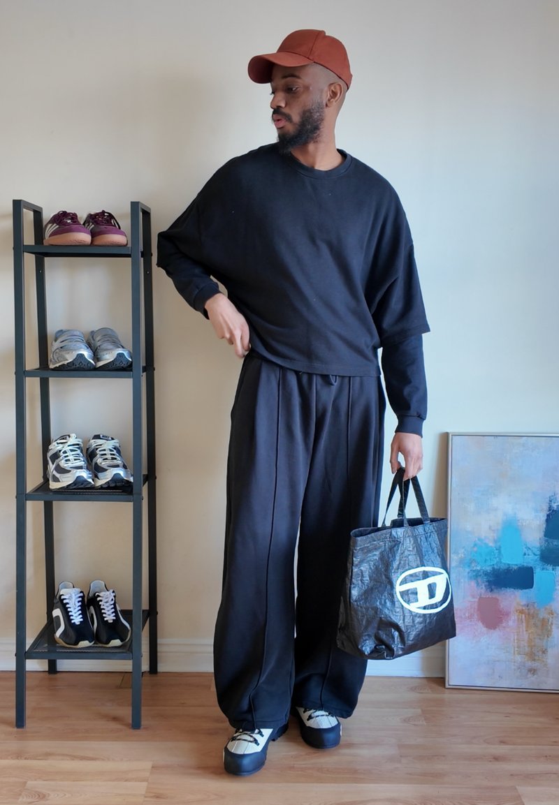 Homme en tenue noire oversize et casquette marron tenant un sac tote noir, debout près d'une étagère à chaussures et d'une peinture abstraite dans une pièce lumineuse.