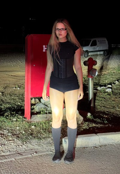 Body de color negro con corsé, tono de piel claro, cabello rubio largo y gafas. Escenario nocturno con una estructura roja y suelo gris.