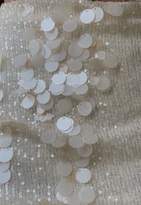 Tissu blanc à paillettes avec des paillettes rondes translucides dispersées de tailles variées. La texture est lisse avec un léger scintillement.