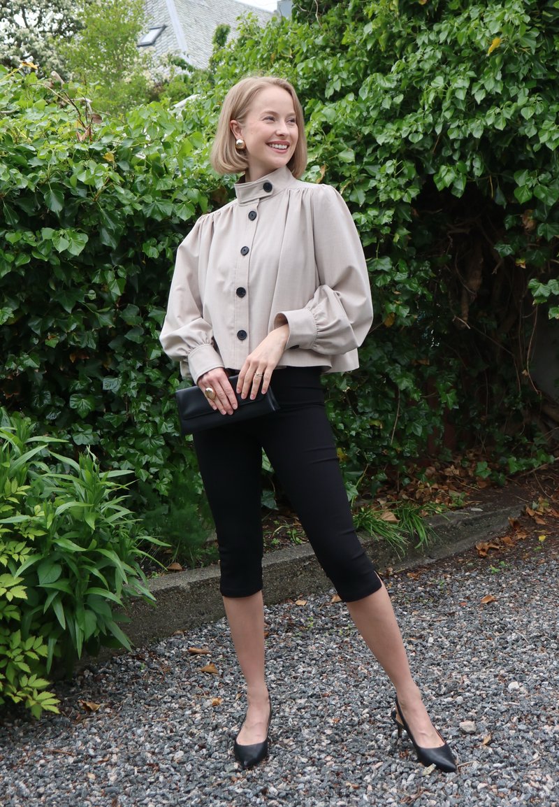 Femme aux cheveux blonds courts portant une veste beige boutonnée, un pantalon noir court, des talons noirs, tenant une pochette noire, debout sur du gravier près de buissons verts.