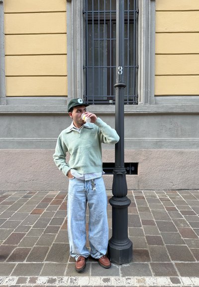 Jersey de punto verde menta, camisa de cuello a rayas debajo, vaqueros holgados azul claros, mocasines marrones, sosteniendo una taza, de pie junto a un farola.