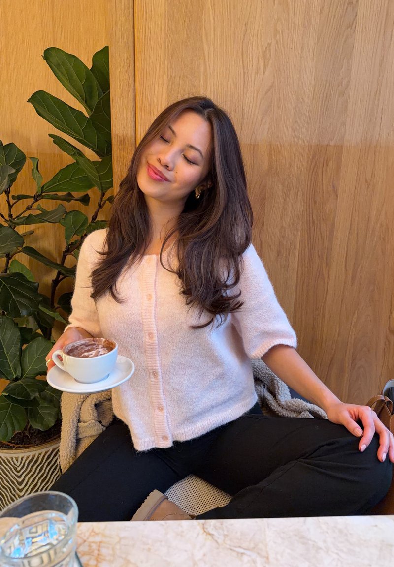 Cardigan rosa chiaro, soffice, con maniche corte e bottoni, indossato con pantaloni neri. Tiene in mano una tazza bianca di cioccolata calda su un piattino.