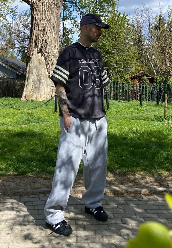 Muž, ktorý má na hlave čiernu čiapku Adidas, čierne tričko Adidas s číslom "03", sivé tepláky a čierne topánky Adidas, stojí na dláždenej ceste blízko trávy a veľkého stromu.