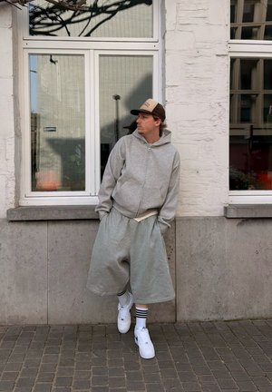 Hombre con sudadera gris con capucha, pantalones cortos grises de gran tamaño, zapatillas blancas y calcetines a rayas, de pie contra una pared de ladrillos blanca con ventanas, mirando de lado.