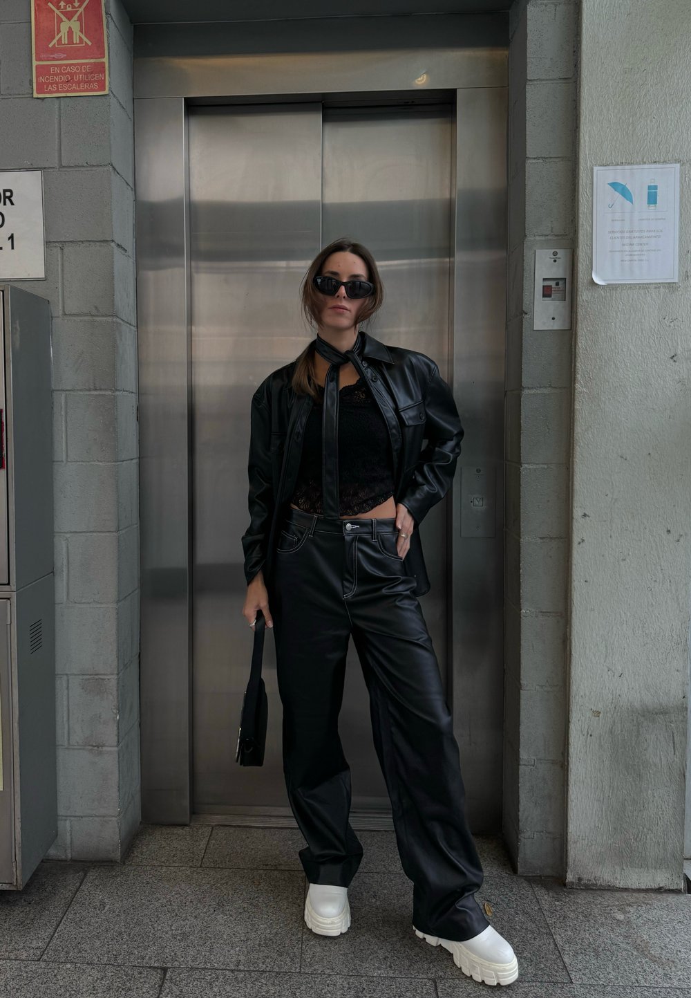 Μαύρο jacket από ψεύτικο δέρμα συνδυασμένο με αντίστοιχο τοπ και χαλαρούς παντελόνια. Λευκά παπούτσια με χοντρή σόλα ολοκληρώνουν το σύνολο. Μαύρα γυαλιά ηλίου και μια μικρή τσάντα περιλαμβάνονται.