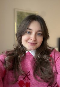 Vaaleanpunainen neule, jossa kirsikkakuvioita, valkoinen kaulus, lainehtiva ruskea tukka, siniset silmät, pehmeä tekstuuri, sisävalaistus, neutraali tausta.