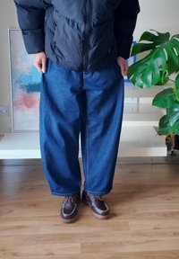 Tmavomodré široké džínsové nohavice, v kombinácii s hnedými koženými topánkami so šnúrkami a dekoratívnym prešívaním. Viditeľná čierna prešívaná bunda.