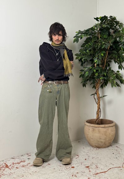 Persona con cabello oscuro y tatuajes, de pie con los brazos cruzados, vistiendo pantalones anchos verdes, sudadera negra, bufanda mostaza, junto a una planta frondosa en maceta.