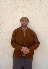 Brown knitted cardigan with large front pockets and a button fastening, worn over a tan shirt, complemented by a brown beanie and glasses.
