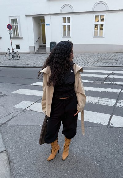 Mujer con cabello largo y rizado que lleva un abrigo beige, ropa negra y botas marrón claro con cordones, parada cerca de un paso de peatones en una calle de la ciudad.