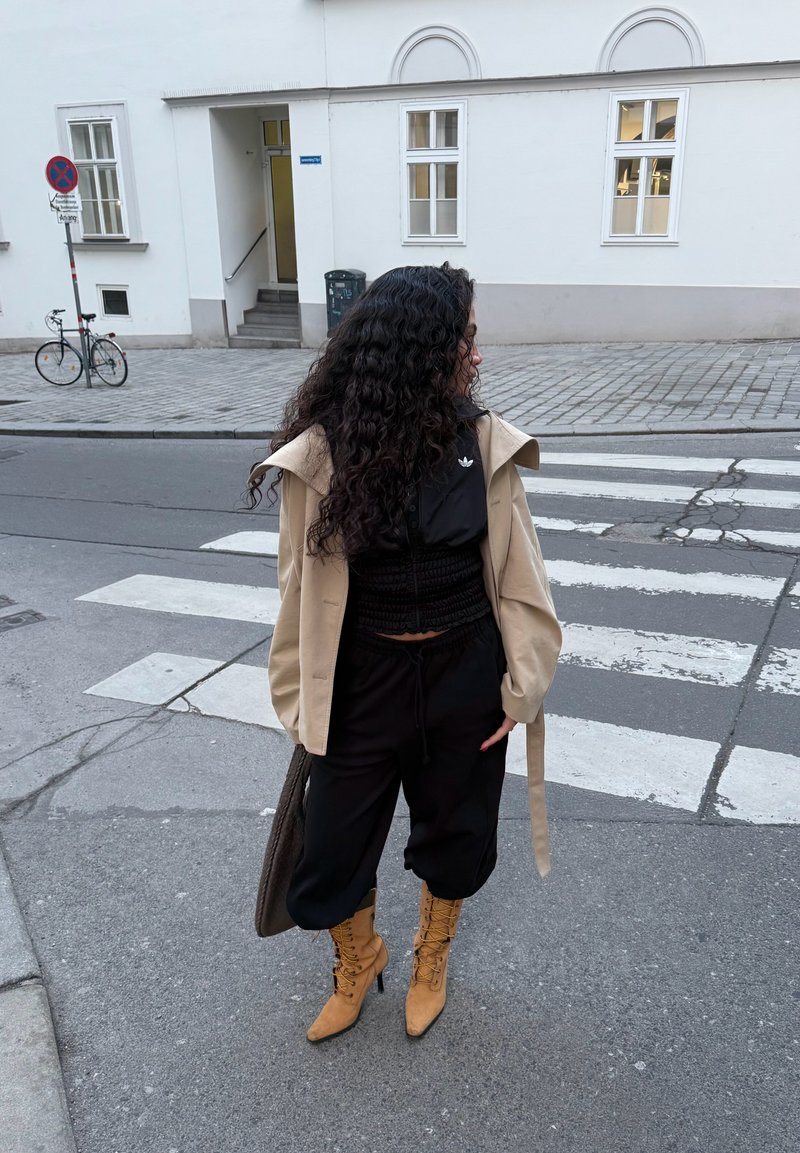Femme aux longs cheveux bouclés portant un manteau beige, une tenue noire et des bottines lacées couleur fauve, debout près d’un passage pour piétons dans une rue de la ville.