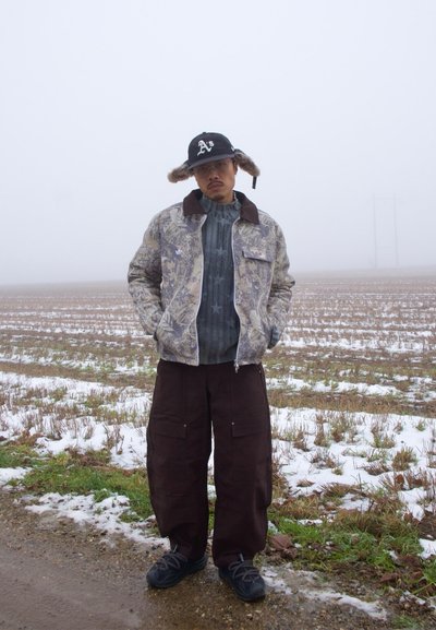 Hombre con chaqueta camuflada, pantalones cargo oscuros, suéter de punto, gorra con ribete de piel y zapatillas de deporte, de pie en un campo nevado bajo un cielo brumoso.